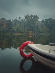 Moody autumn background with a boat on the calm lake water with a vibrant red lifeline ring. Colorful fall trees reflecting on the pond surface in a calm foggy day in the park. Cloudy gloomy sky.