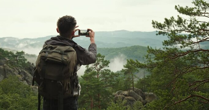 A man is standing on top of a cliff and taking pictures of nature. He is carrying a large backpack. Hiking in the mountains. Fog in the mountains. 4K DCI