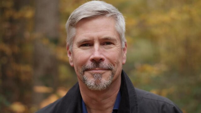 Portrait of a smiling handsome middle aged man in nature, slow motion. Mature caucasian man showing real emotions being happy, confident and likable. Attractive autumn forest background.