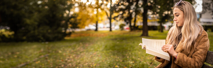 Panorama young woman sitting in the park on a bench and reading a book. In the background are beautiful autumn colors.