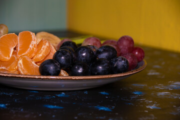 Fruits, grapes, tangerines, bananas, kiwi in a blue porcelain plate. black wooden floor, fruit plate