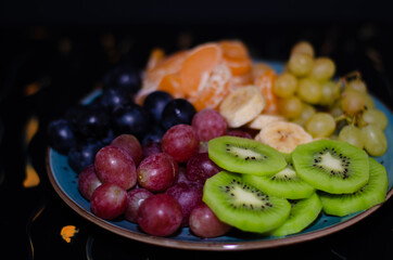 Fruits, grapes, tangerines, bananas, kiwi in a blue porcelain plate. black wooden floor, fruit plate