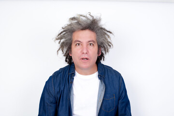 Handsome middle aged Caucasian man with afro gray hair, against white wall,  having stunned and shocked look, with mouth open and jaw dropped exclaiming: Wow, I can't believe this. Surprise and shock