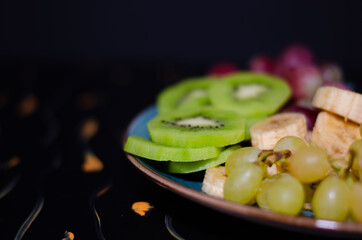 Fruits, grapes, tangerines, bananas, kiwi in a blue porcelain plate. black wooden floor, fruit plate