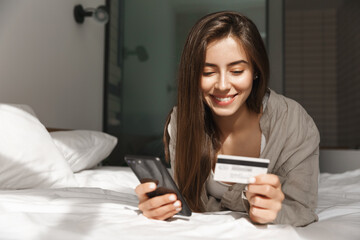 Fototapeta na wymiar Young woman with smartphone and credit card lying in bed at home, shopping online