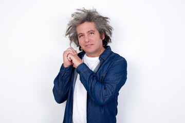 Dreamy Handsome middle aged Caucasian man with afro gray hair, against white wall,  with pleasant expression, closes eyes, keeps hands crossed near face, thinks about something pleasant