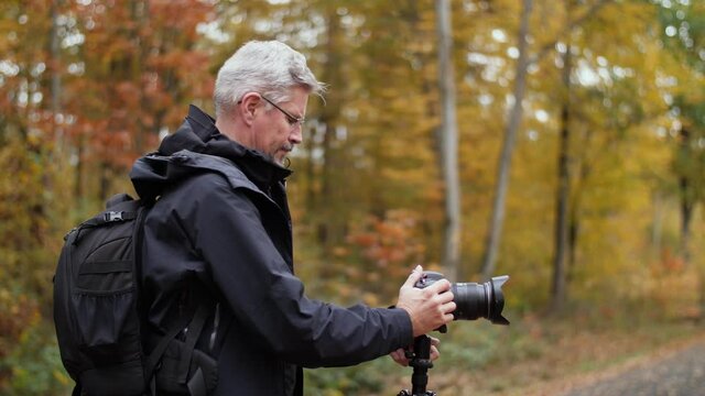 Mature photographer adjusts settings on a DSLR camera in nature in late autumn. Male Caucasian takes pictures outdoors with a professional camera. Beautiful forest background. Using a tripod.