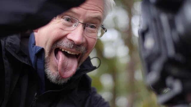 Joyful adult photographer makes funny faces while looking at his camera. Male Caucasian stands in nature with a beautiful forest background. Using a tripod.
