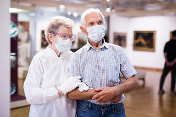 mature couple in mask protecting against covid examines paintings on display in hall of art museum