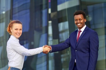 Two happy smiling people: businessman and businesswoman are shaking hands, greeting. White European young woman and black African Afro American man in formal suit outdoors, looking at camera