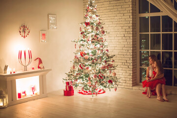 Merry Christmas and Happy Holidays. Mom and daughter with a Christmas tree indoors at night. The evening before Christmas. Portrait of a loving family nearby. High quality photo.