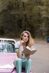 pensive woman sitting on hood of cabriolet and reading book on blurred background, banner