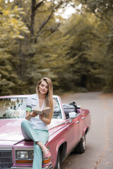 Obraz na płótnie Canvas young smiling woman sitting on hood of vintage cabriolet and reading book