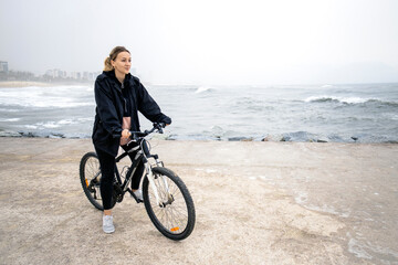 Fototapeta premium Side view a young beautiful blond woman riding on bicycle on the beach with sea on background on rainy and cloudy day. Space for text