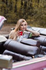 smiling woman holding hand near face and looking away while sitting in cabriolet with cup of coffee...