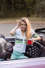 smiling woman touching hair while holding cup of coffee in vintage cabriolet