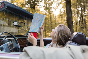 young woman looking at map while sitting in convertible car on blurred foreground