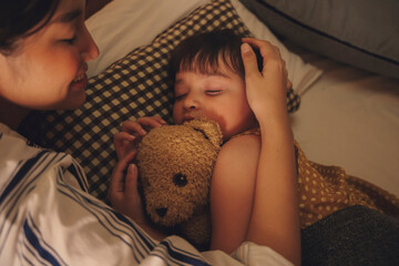 Mother watching her child sleep and hug teddy bear on a bed at night.
