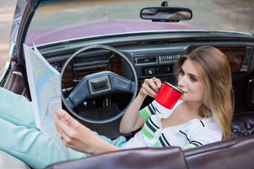 young woman drinking coffee and looking at road atlas in cabriolet on blurred foreground