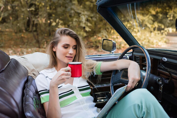 young woman looking at road map while sitting in cabriolet with cup of coffee