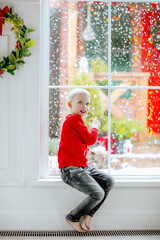 Boy in winter sweater and jeans sitting on a big window with snowing background.