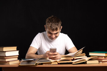 A YOUNG MAN of European appearance sits at a table with books and looks into a smartphone. DISTANCE LEARNING in quarantine. an accountant works in an office against a black wall.