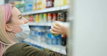 Fair haired lady with pink strand wearing blue medical mask chooses beverage can from rack in supermarket close view slow motion