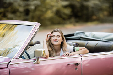 happy young woman holding hand near face while sitting in cabriolet and looking at camera