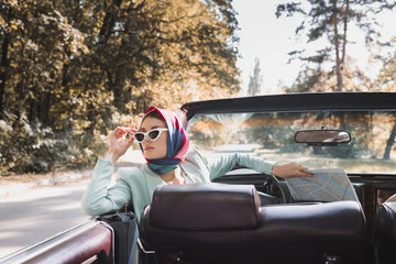 Stylish woman touching sunglasses while holding map in retro car on blurred foreground