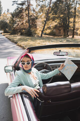 Stylish woman holding map while sitting on driver seat of cabriolet car on road