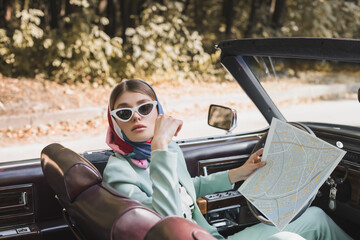 Fashionable woman in sunglasses holding map on driver seat of car on blurred foreground