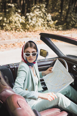 Elegant woman with map looking away in retro car on blurred foreground during trip