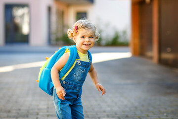 Cute little adorable toddler girl on her first day going to playschool. Healthy beautiful baby walking to nursery school and kindergarten. Happy child with backpack on the city street, outdoors.