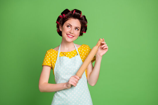 Photo of charming cheerful lady curlers hairdo housewife hold rolling pin kitchen look up empty space dreamy thinking wear pinup dotted dress apron isolated green color background