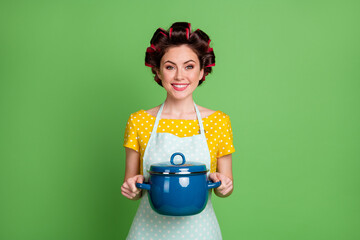 Photo of positive girl house wife hold saucepan enjoy prepare organic supper wear yellow dotted t-shirt hair rollers isolated over green color background