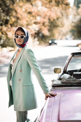 trendy woman in kerchief and sunglasses standing on road near cabriolet