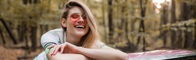 excited young woman in sunglasses looking at camera while leaning on car, banner