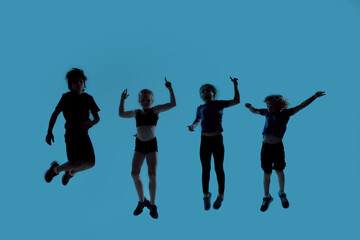 Fototapeta na wymiar Silhouetted full length shot of four little sportive kids looking joyful while posing, jumping isolated over blue background