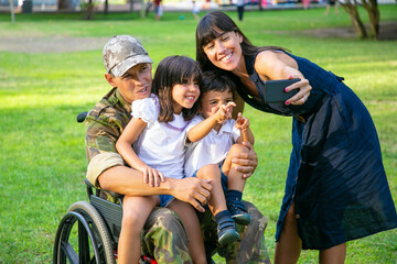 Happy disabled retired military man holding kids in arms while his wife taking family selfie on cell phone. Veteran of war or returning home concept - Powered by Adobe