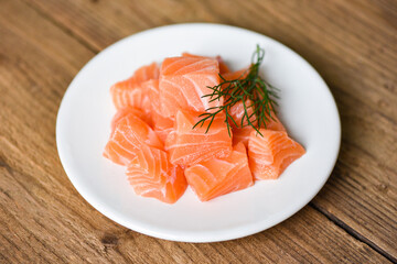 Raw salmon filet cube with herbs and spices on white plate on wooden background