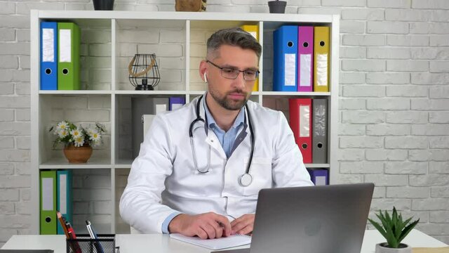 Man Doctor Wears White Medical Coat Wireless Earphones Consults Patient Remote Online Video Call Laptop Webcam. Male Nurse In Hospital Office Distant Listen Speak Client Uses Virtual Chat Computer App