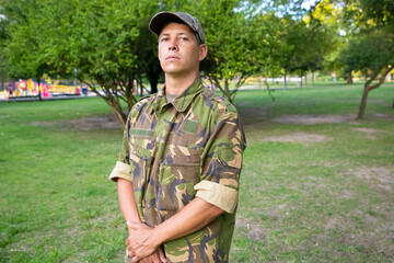 Portrait of guard in park. Serious man in military camouflage uniform standing in park, looking at...