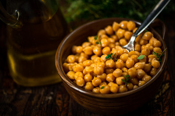 Boiled chickpeas in bowl with spoon, carafe with olive oil, herb in dark background.