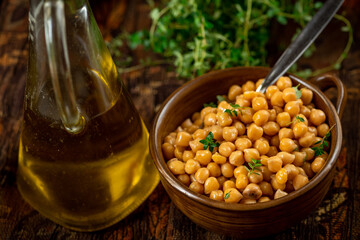 Chickpeas in bowl with spoon, glass carafe with olive oil, thyme herb on dark table.