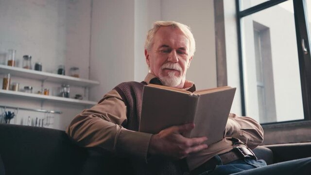 Low angle view of smiling elderly man reading book while sitting at sofa at home