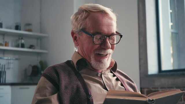 Smiling pensioner in eyeglasses reading book with blurred kitchen on background