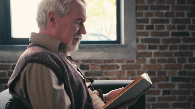 Side view of positive senior man reading book and laughing, sitting near window
