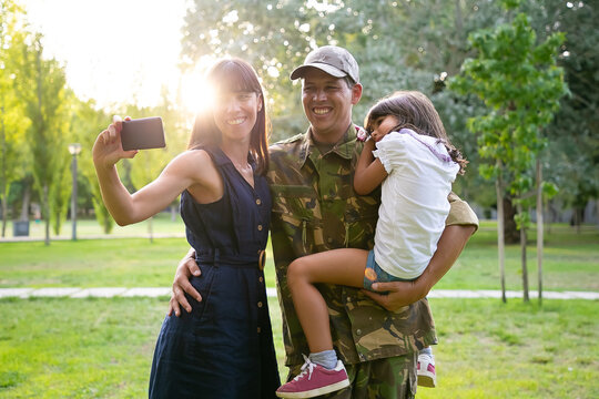 Happy military couple of parents and little daughter taking selfie on cellphone in city park. Medium shot, sunshine. Family reunion or returning home concept