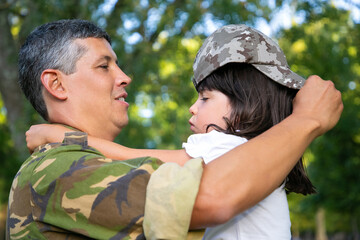 Happy dad holding daughter in arms, dressing girl in his camouflage cap. Side view. Family reunion or returning home concept