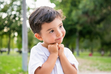 Cheerful sweet little boy standing and posing in summer park, leaning chin on hands, smiling and looking away. Closeup shot. Childhood concept